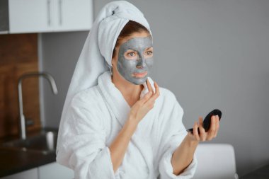 Beyaz bornoz ve havlu giymiş güzel beyaz kadın güzellik maskesi kullanıyor.