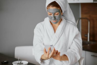 Bornoz ve havlu giymiş çekici genç bir bayan yüz maskesi ve akıllı telefon kullanıyor.
