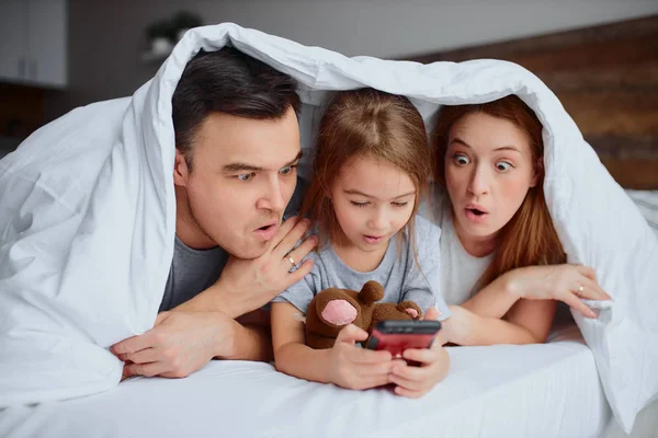 Emotionele ouders en dochter kijken video op smartphone op bed — Stockfoto