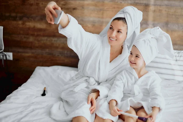 Hermosa mujer y niño pequeño tomando selfie usando albornoz en la cama —  Fotos de Stock