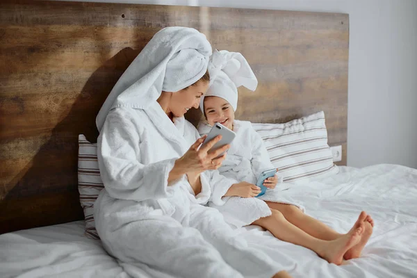 Atractiva mujer con hija se acuesta en la cama con teléfono móvil —  Fotos de Stock