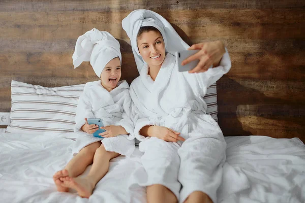 Hermosa mujer y niño pequeño tomando selfie usando albornoz en la cama —  Fotos de Stock