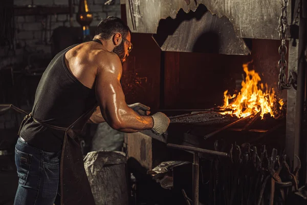 Giovane fabbro lavorare a temperatura calda durante il riscaldamento pezzo di acciaio in forno — Foto Stock