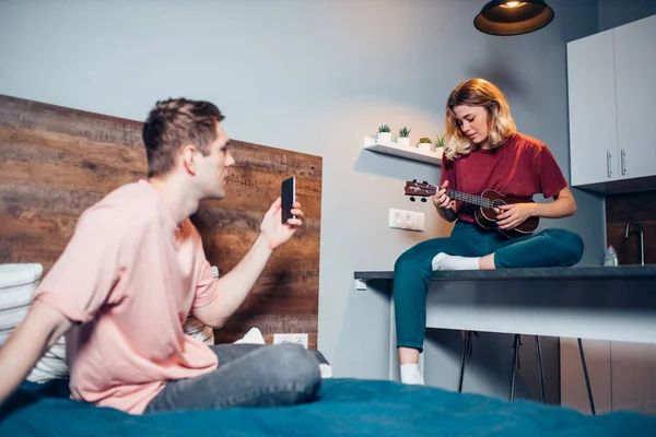 Atractiva mujer jugando ukelele en casa, mientras que su amigo tomar fotos o grabar vídeo — Foto de Stock