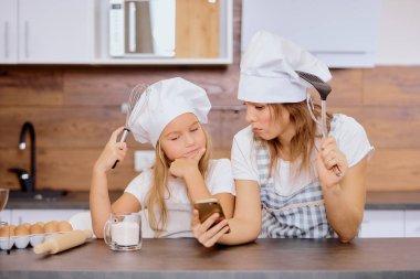 Genç anne ve kız mutfakta birlikte yemek yaparken akıllı telefon kullanıyor.