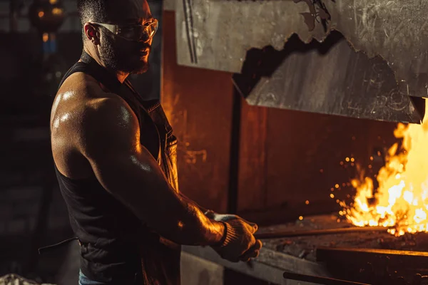 Joven herrero caucásico a temperatura caliente mientras calienta la pieza de acero en el horno — Foto de Stock