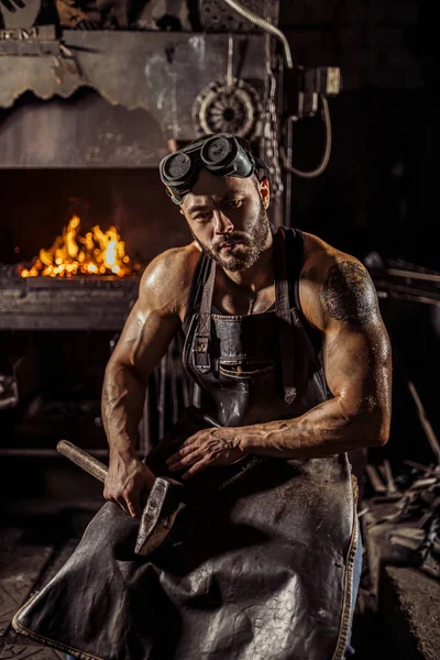 Portrait of young caucasian blacksmith man in leather black uniform works in smithy — Stock Photo, Image