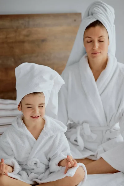 Retrato del niño y su madre meditando con los ojos cerrados en la pose de loto en la cama en casa —  Fotos de Stock