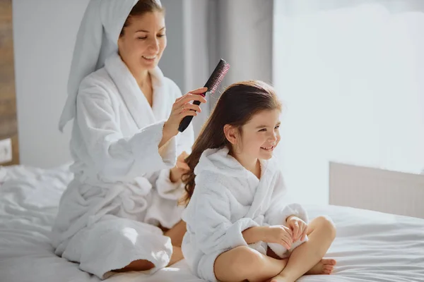 Sonriente familia amigable de madre e hija peinando el pelo en la cama —  Fotos de Stock