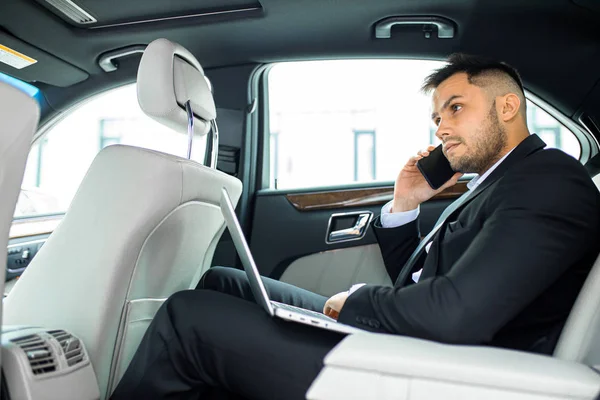 Junger Geschäftsmann arbeitet am Laptop und spricht im Auto mit dem Smartphone — Stockfoto