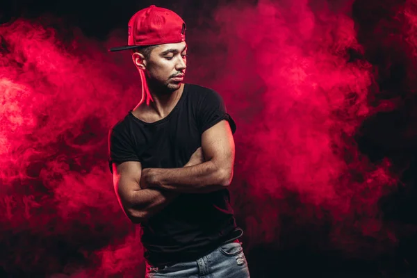 Retrato de homem vigoroso agradável vestindo t-shirt preta, jeans e boné isolado sobre fundo fumegante vermelho — Fotografia de Stock