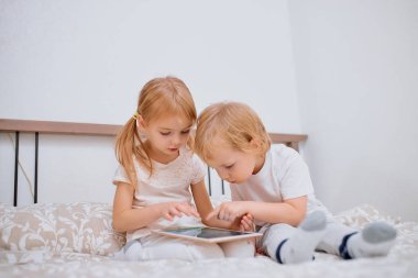 Küçük beyaz çocuklar yatakta otururken tablet kullanıyorlar.