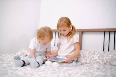 Küçük beyaz çocuklar yatakta otururken tablet kullanıyorlar.