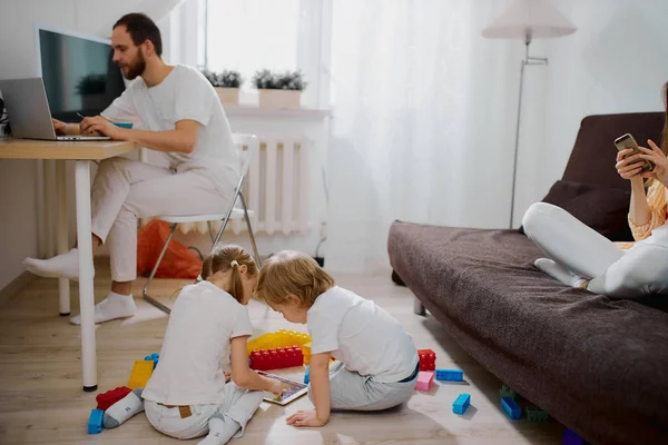 Niños jugando juntos en el suelo mientras los padres jóvenes se relajan en casa —  Fotos de Stock