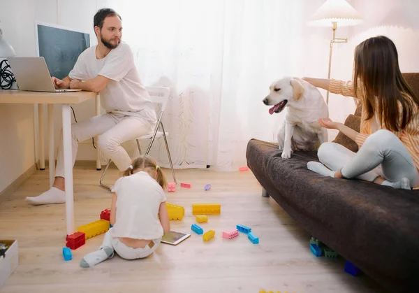 Dzieci bawią się razem na podłodze, podczas gdy młodzi rodzice relaksują się w domu — Zdjęcie stockowe