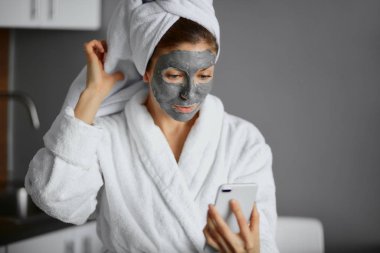 Bornoz ve havlu giymiş çekici genç bir bayan yüz maskesi ve akıllı telefon kullanıyor.