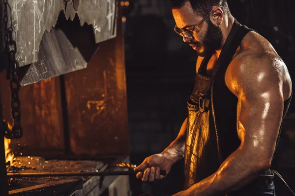El herrero joven trabaja a temperatura caliente mientras calienta la pieza de acero en el horno — Foto de Stock