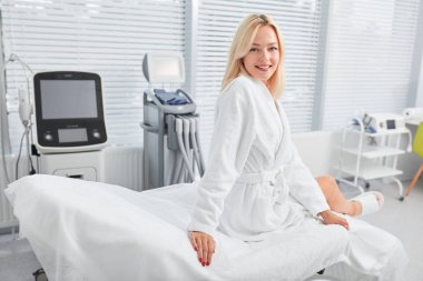 Güzel, genç, neşeli, beyaz bornozlu ve sandalyede oturan sarışın kadın.