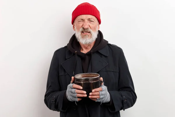 Viejo vagabundo con barba gris con abrigo negro sosteniendo una lata de hierro por dinero — Foto de Stock
