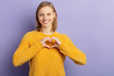 Genç, gülümseyen bir kadın, mor arka planda izole edilmiş bir şekilde kameraya kalbini gösteriyor.