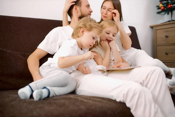 Attractive happy, family consisted of mother, father, son and daughter sitting on sofa at home with tablet — Stock Photo, Image