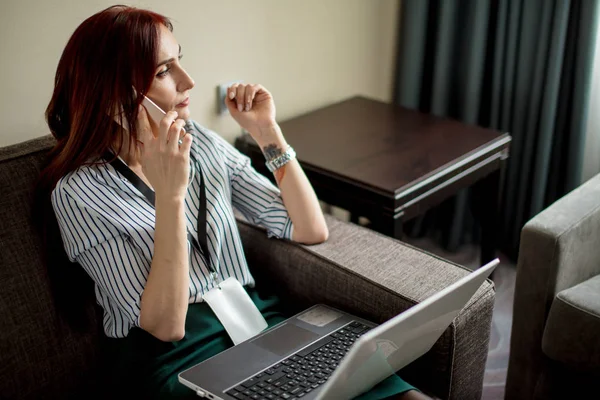 Kaukasische Rotschopf elegante Frau mit Telefon und Laptop sitzt auf Sofa im Hotel — Stockfoto