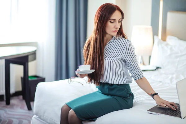 Donna dai capelli rossi in abito formale che prende il caffè e lavora su un computer portatile a letto . — Foto Stock