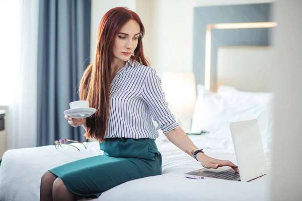 Rothaarige Frau in offizieller Kleidung beim Kaffee und bei der Arbeit an einem Laptop im Bett. — Stockfoto