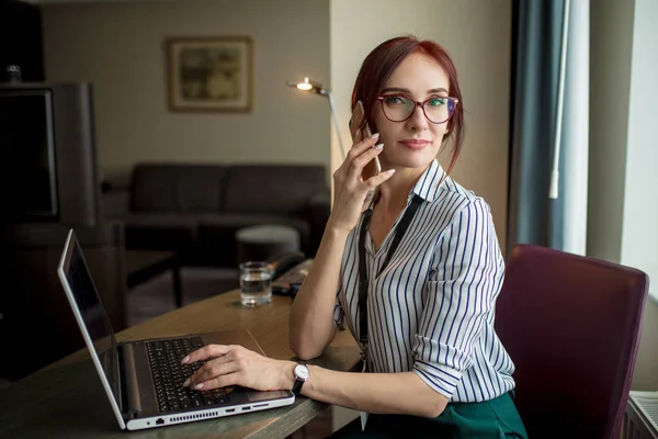 Zrzavý žena sedí u stolu a práci na notebooku a mluvit na telefonu — Stock fotografie