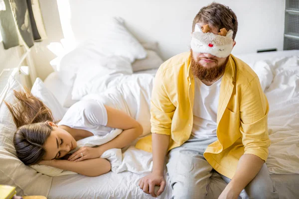 Νυσταγμένος άντρας με μάσκα κάθεται στο κρεβάτι δίπλα σε κοιμισμένη γυναίκα — Φωτογραφία Αρχείου