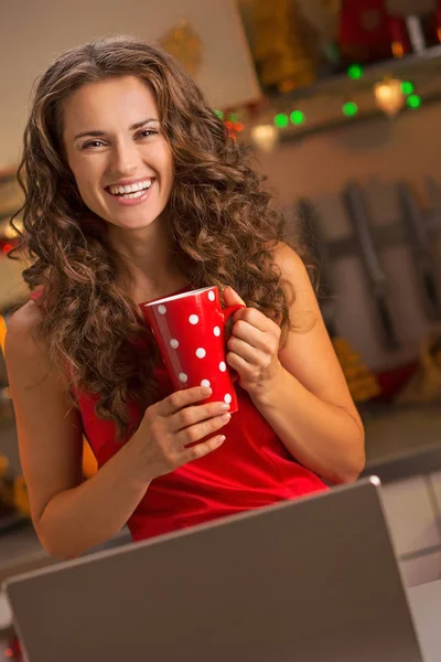 Glückliche junge Hausfrau mit Tasse heißer Schokolade mit Laptop in — Stockfoto