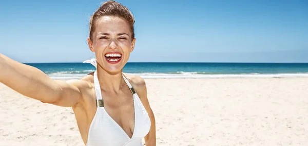 Mujer sonriente en traje de baño blanco tomando selfie en la playa de arena —  Fotos de Stock