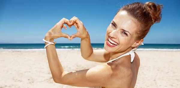 Mujer en traje de baño mostrando las manos en forma de corazón en la playa de arena —  Fotos de Stock