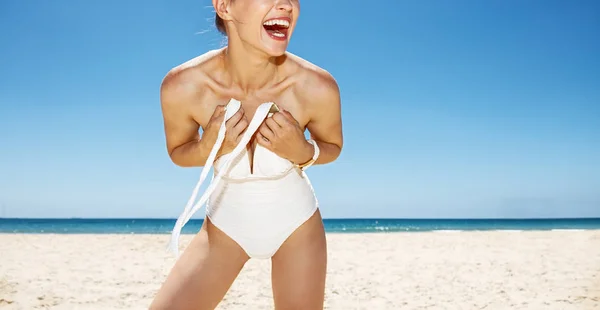 Mujer sonriente atando traje de baño blanco en la playa de arena en el día soleado — Foto de Stock