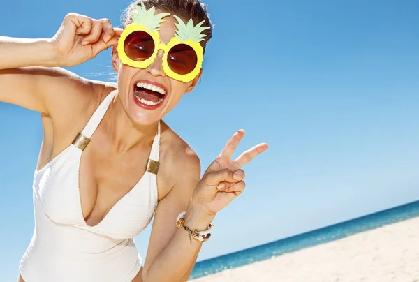 Woman in pineapple glasses showing victory gesture at beach — Stock Photo, Image