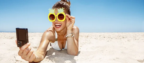 Mulher sorridente em óculos de abacaxi tomando selfie na praia de areia — Fotografia de Stock
