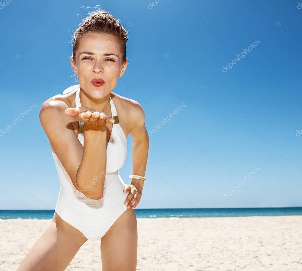 Smiling woman in white swimsuit blowing air kiss at sandy beach