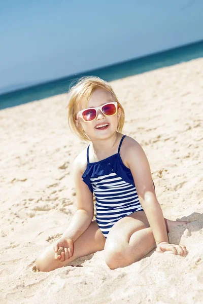 Glückliches Mädchen im gestreiften Badeanzug spielt mit Sand am weißen Strand — Stockfoto