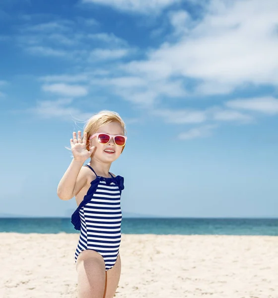 Glückliches Mädchen im gestreiften Badeanzug an einem weißen Strand beim Händewinken — Stockfoto