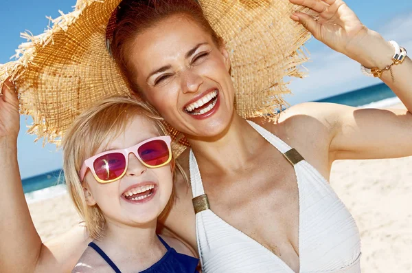 Smilende mor og datter under stor halm hat på sandstrand - Stock-foto