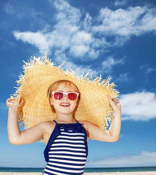 Happy girl in striped swimsuit and big straw hat on white beach — Stock Photo, Image