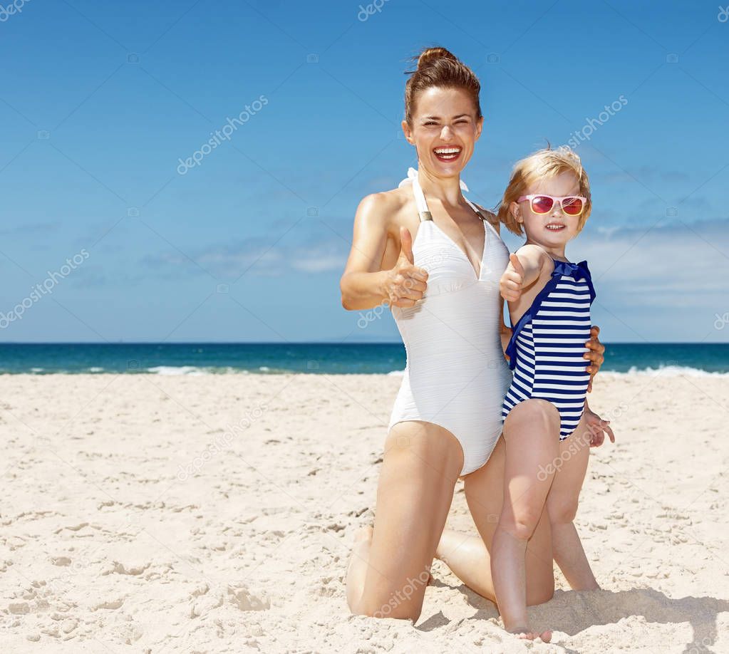 Happy mother and child in swimsuits at beach showing thumbs up
