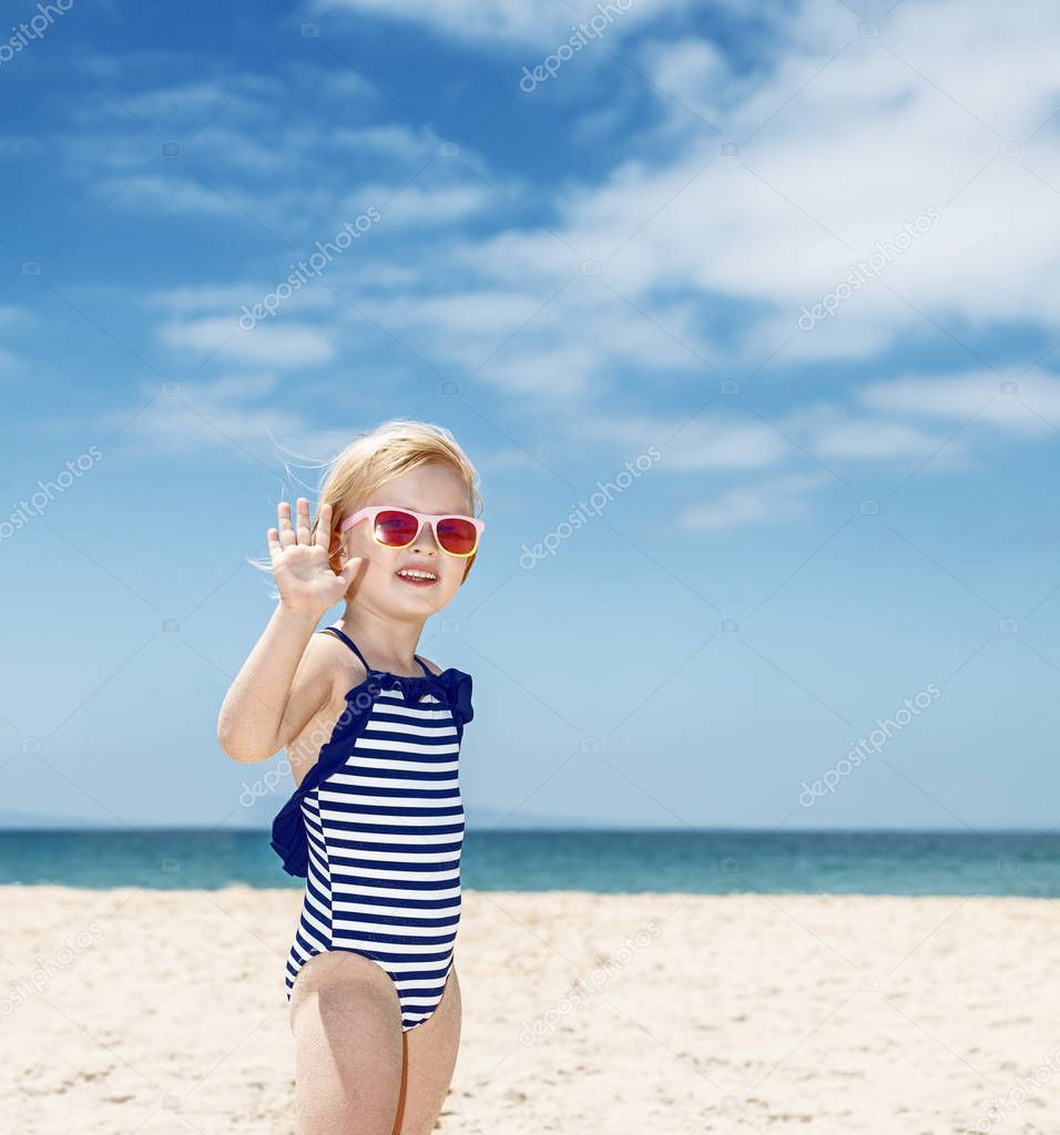 Happy girl in striped swimsuit on a white beach handwaving