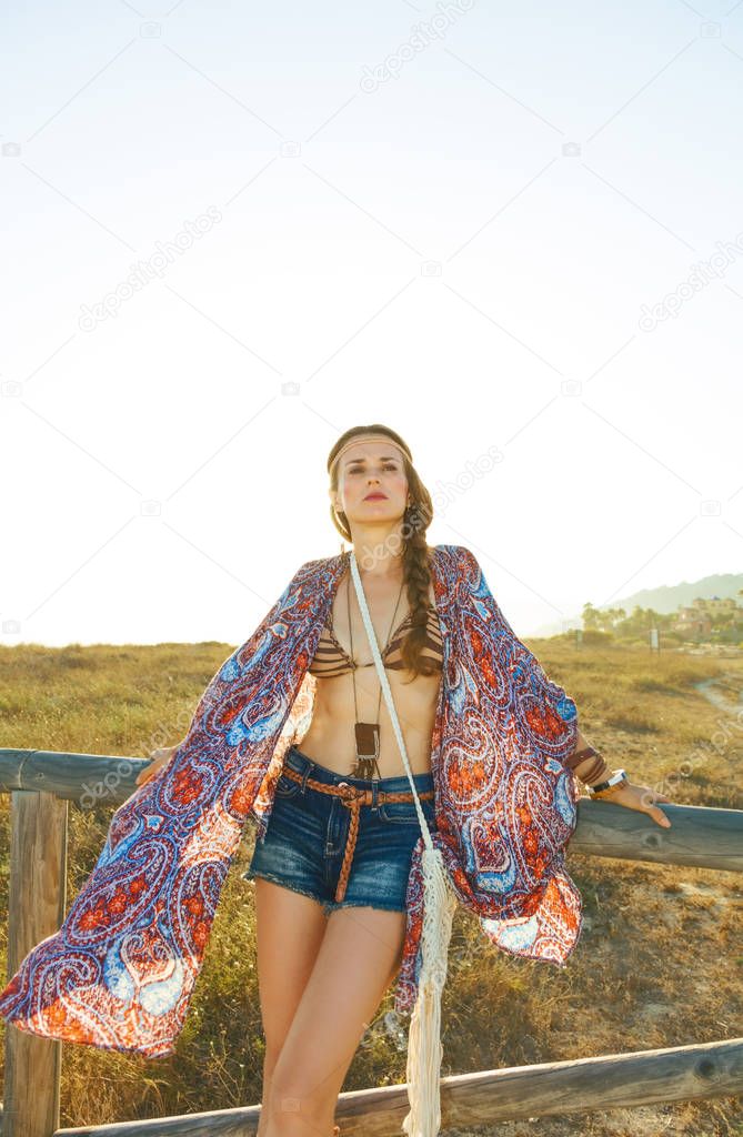 Bohemian vibe vacation. Portrait of young bohemian girl in jeans shorts and cape outdoors in the summer evening looking into the distance