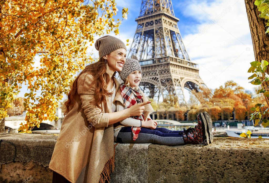 Portrait of smiling mother and child travelers on embankment near Eiffel tower in Paris, France pointing on something