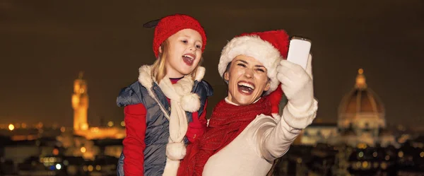 旅行充满灵感在圣诞节的时候在佛罗伦萨 微笑的年轻的母亲和儿童游客在圣诞帽在佛罗伦萨的全景视图以与智能手机拍照 — 图库照片
