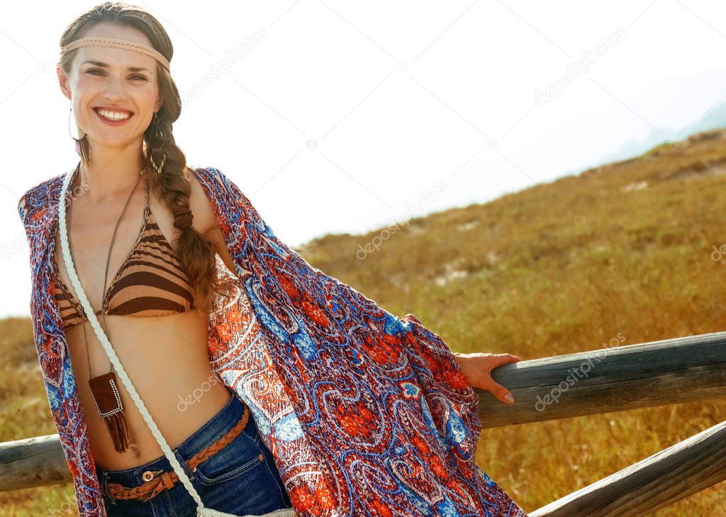 Bohemian vibe vacation. smiling trendy boho girl in jeans shorts and cape outdoors in the summer evening near a fence