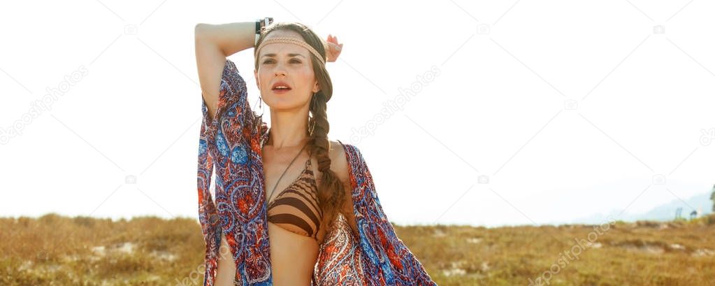 Bohemian vibe vacation. Portrait of modern free spirit girl in jeans shorts and cape outdoors in the summer evening looking into the distance near a fence