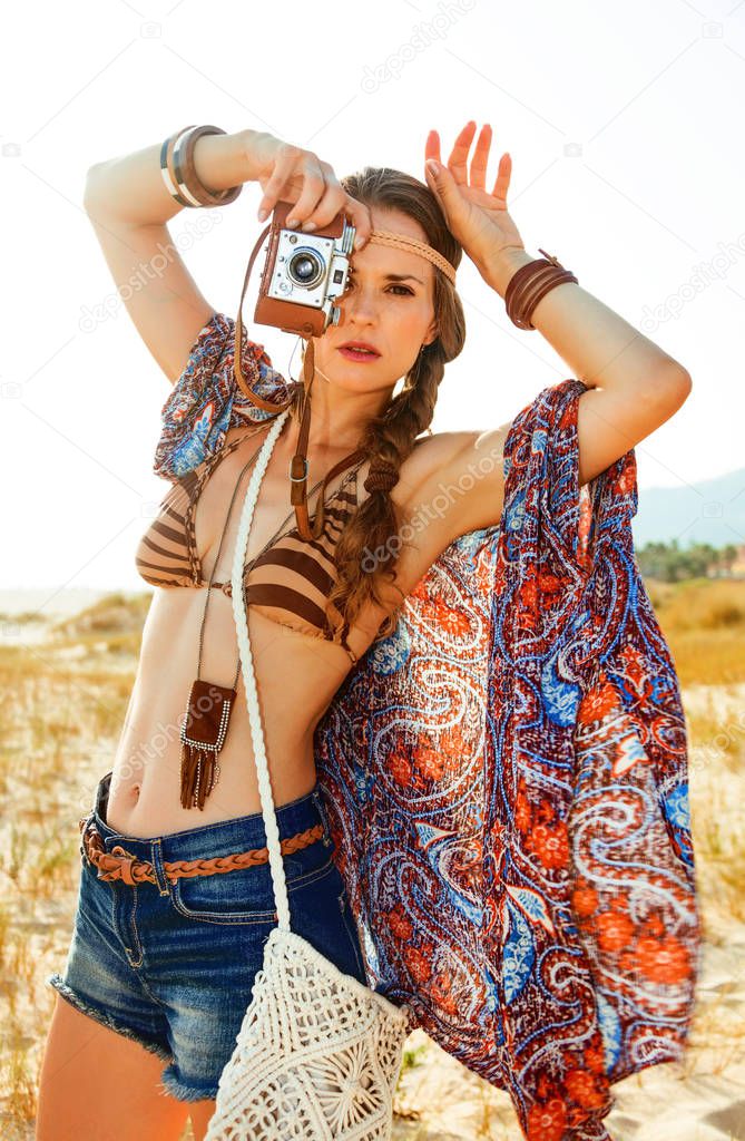 Bohemian vibe vacation. Portrait of trendy hippie in jeans shorts and cape outdoors in the summer evening with retro photo camera