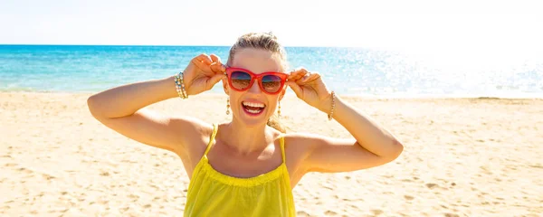 Portrait of happy modern woman in colorful dress on the seashore holding sunglasses